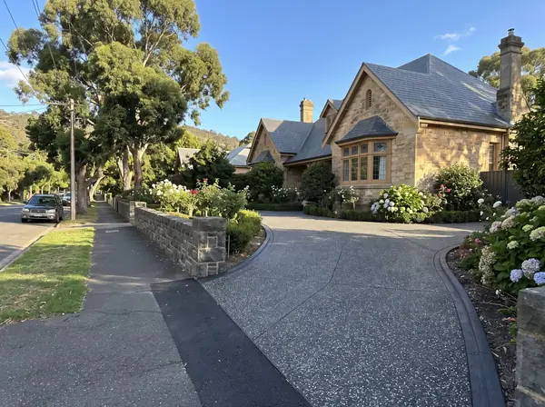 A new charcoal exposed aggregate driveway sweeping up past a large sandstone home under huge gum trees in Burnside