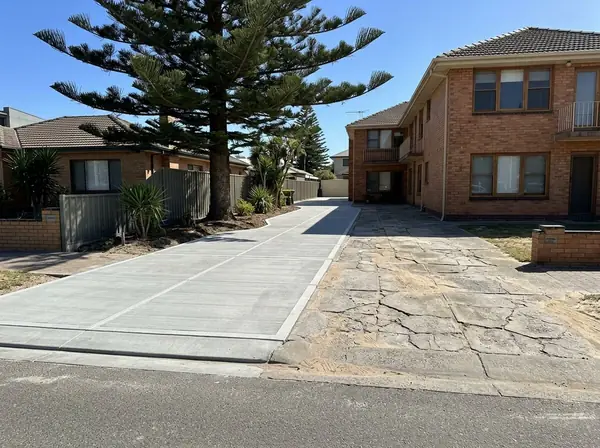 An old cracked, salt-stained driveway at a 1960s unit block with sand blown across it beside a neighbour's new pale exposed aggregate driveway, Glenelg
