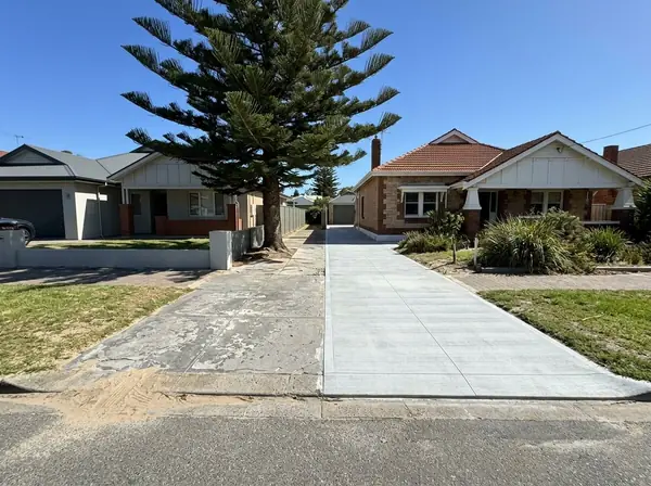 An old flaking, salt-stained bungalow driveway with sand blown across it beside a neighbour's new pale driveway, Henley Beach