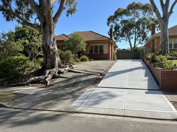 An old cracked sloping driveway lifted by a gum root beside a 1930s bungalow, next to a neighbour's new exposed aggregate driveway with a strip drain, Mitcham