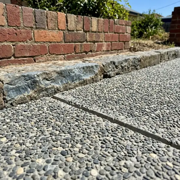 Close up of a new exposed aggregate driveway edge meeting an old bluestone kerb and a red brick wall